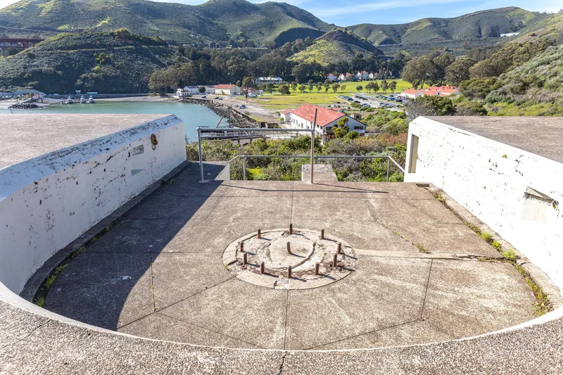 Aerial view of a seaside battery with hills and village, sunlit under a clear sky.