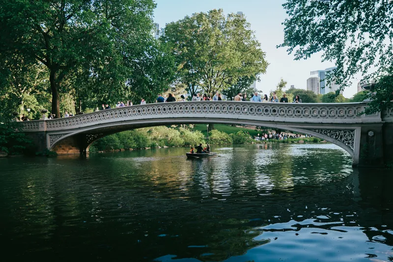 A picturesque scene of Bow Bridge with people and a boat in Central Park, New York City.