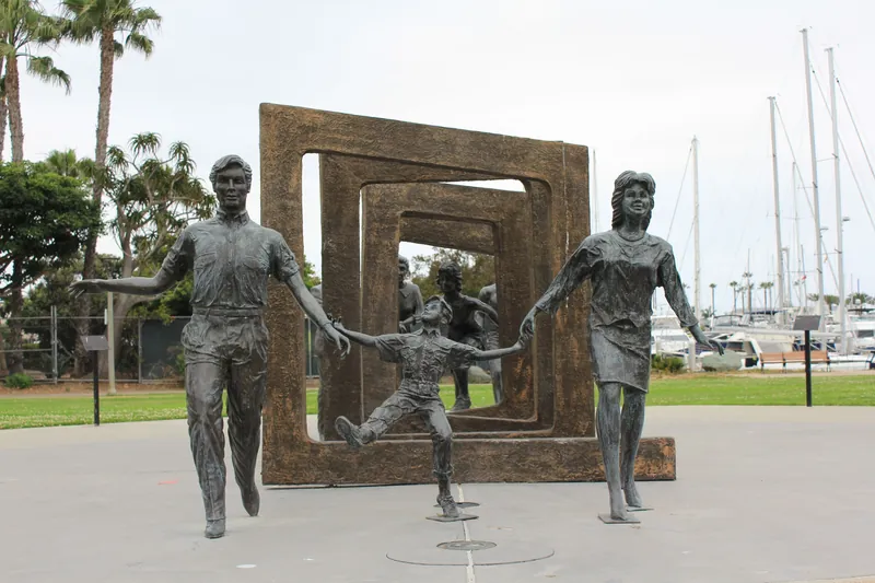 Bronze statues of a family in a park, framed by geometric sculptures and marina backdrop.