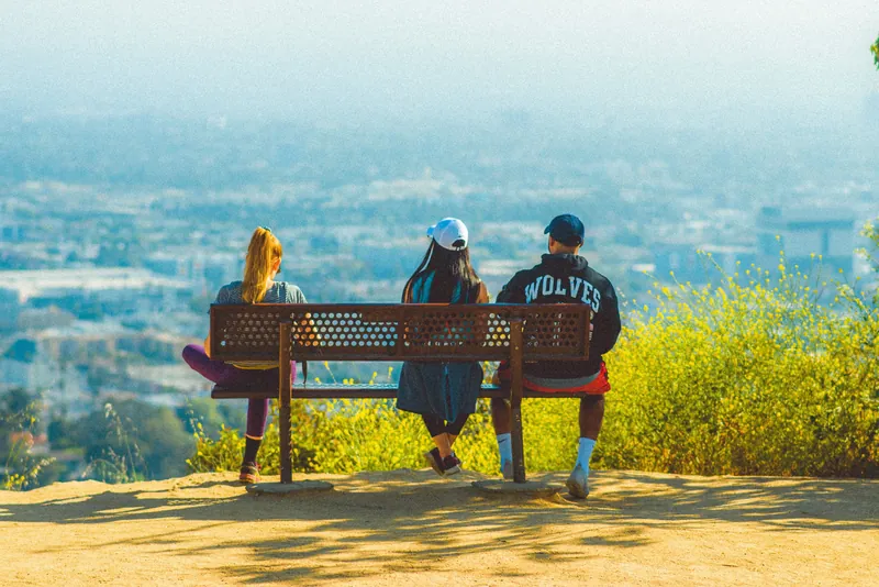 Three young adults sit on a bench enjoying a panoramic view of the city on a sunny day.
