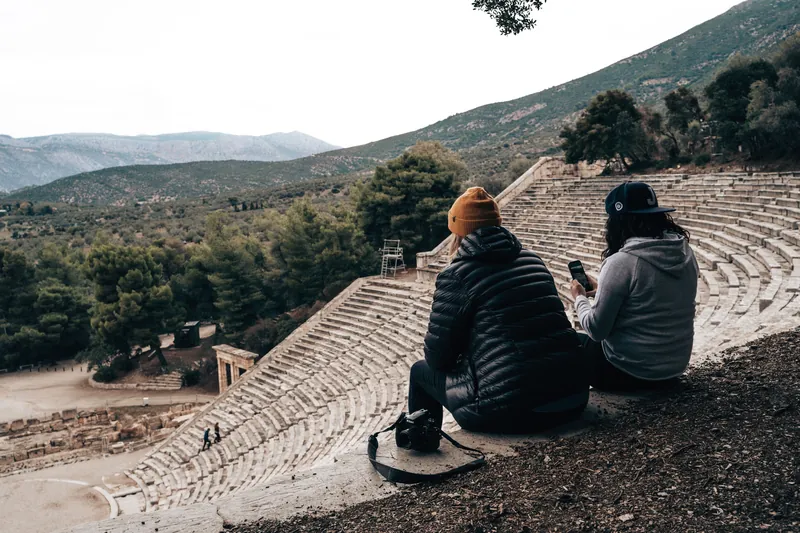 Two women sitting in an ancient Greek amphitheater with scenic mountain views.