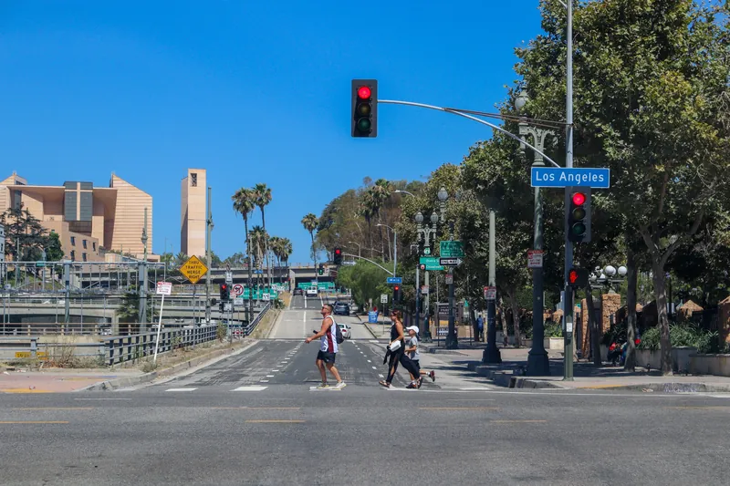 Pedestrians crossing a street in downtown Los Angeles under sunny blue skies with iconic buildings in view.