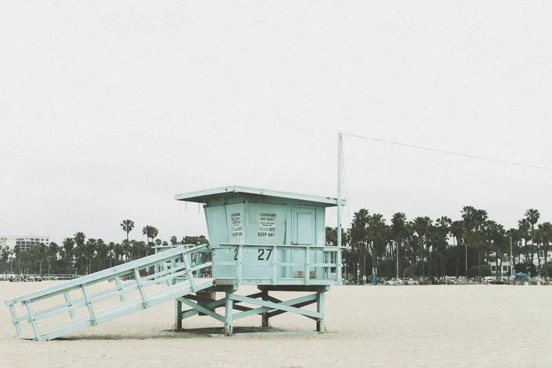 A serene view of a lifeguard tower on a Los Angeles beach surrounded by palm trees under an overcast sky.