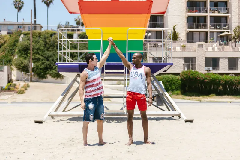 Two men celebrate LGBT pride at a colorful lifeguard station on a sunny beach.