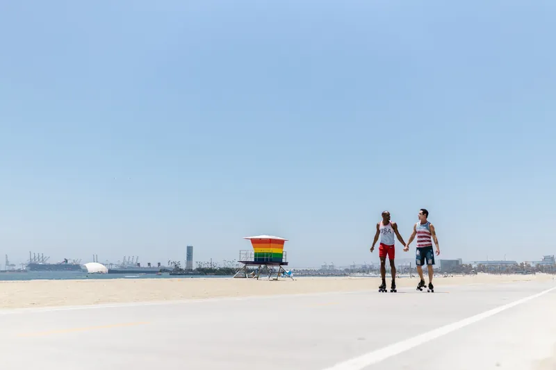 Two men holding hands while roller skating on a sunny beach, featuring a rainbow-colored lifeguard tower.