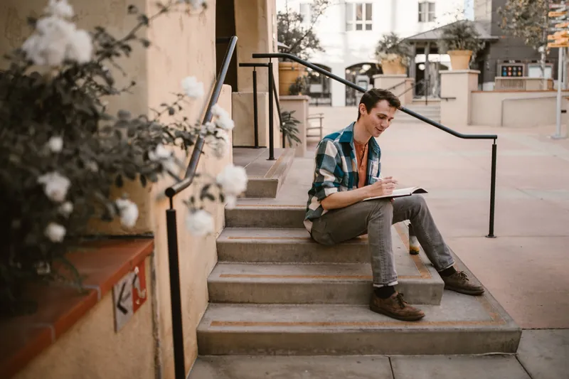 A young man in plaid shirt and jeans writing in a notebook while sitting on concrete steps.
