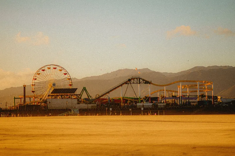Scenic view of Santa Monica Pier with ferris wheel and roller coaster against mountain backdrop at dusk.