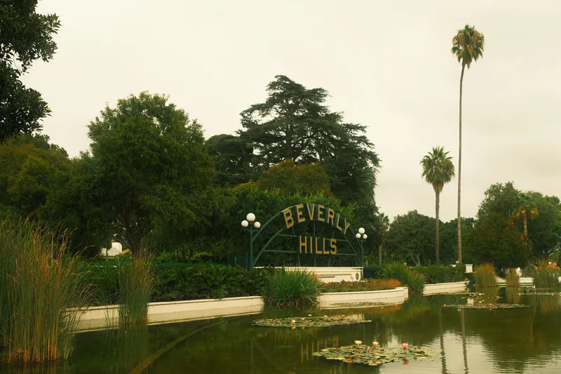 Tranquil Beverly Hills sign by a lush pond with palm trees on a cloudy day.