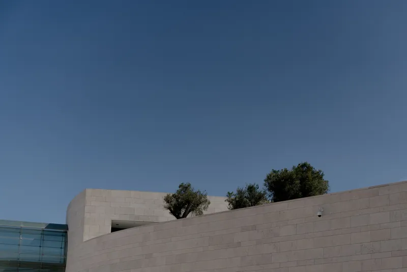 A modern building with sleek lines against a clear blue sky, featuring textured concrete walls and trees.