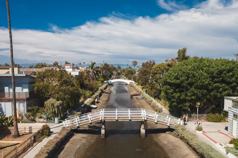 A picturesque view of the Venice Canals featuring a white pedestrian bridge and lush greenery under a blue sky.