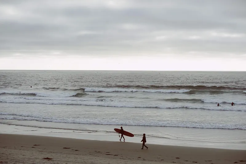 Surfer strolling along San Diego beach under a cloudy sky, capturing ocean waves at sunset.